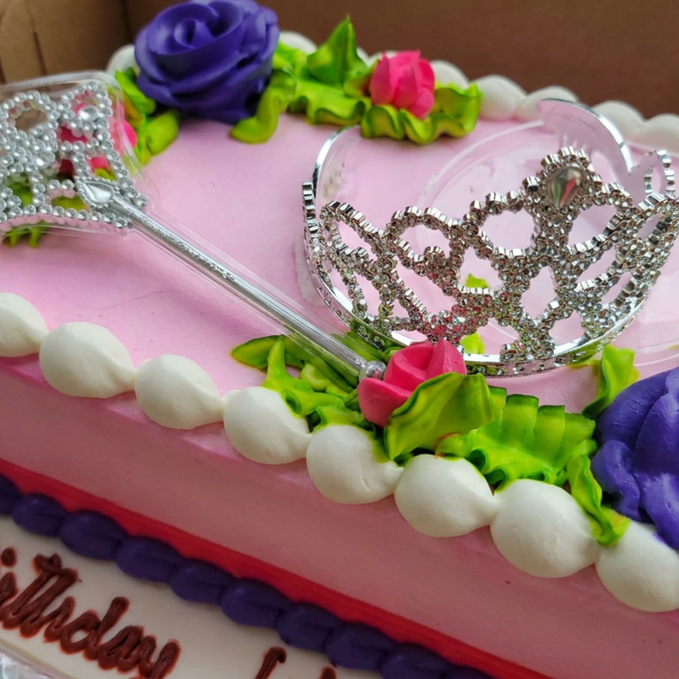 Pink birthday cake decorated with colorful buttercream flowers, white piping, a silver tiara, and “Happy Birthday” lettering.