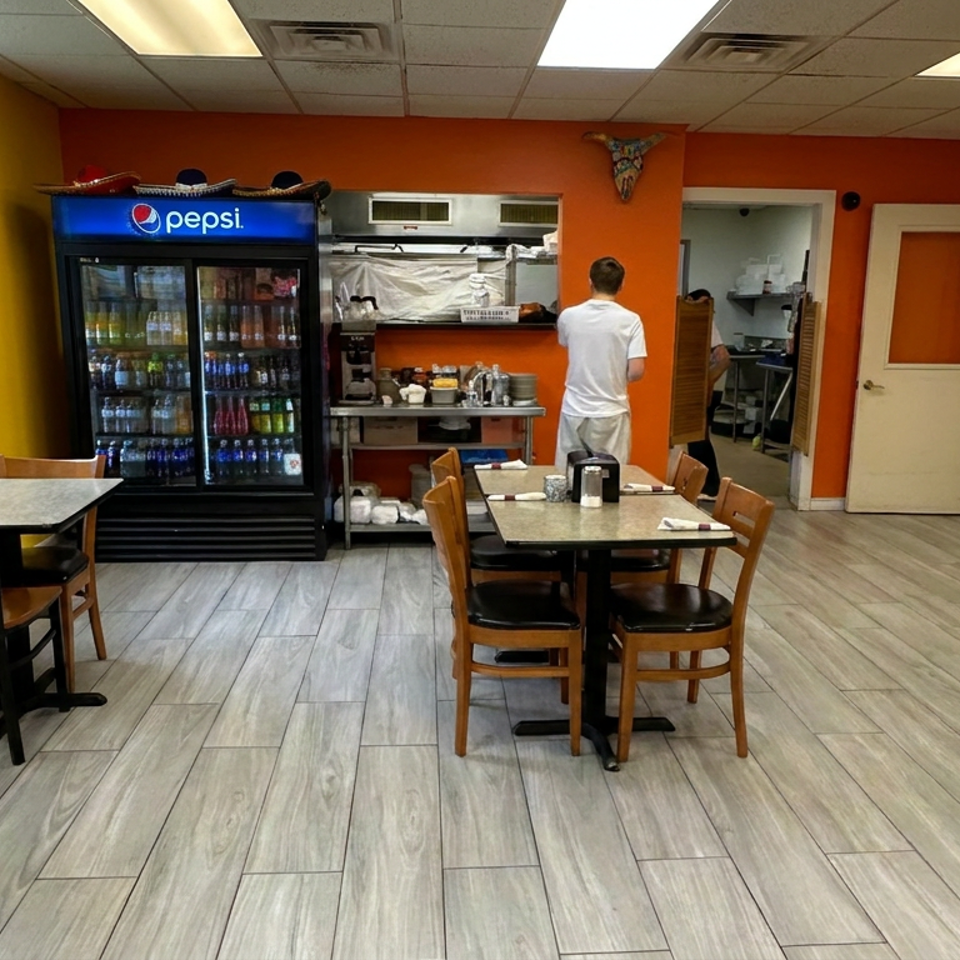 Bright casual restaurant dining room with wooden tables, chairs, beverage cooler, service counter, and a worker in the kitchen area.